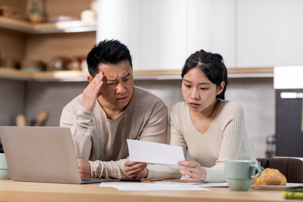 Upset korean husband and wife sitting at table in front of computer, reading letter from bank, shocked with their debts, kitchen interior, copy space. Financial hangover during 2022 crisis - Φωτογραφία, εικόνα