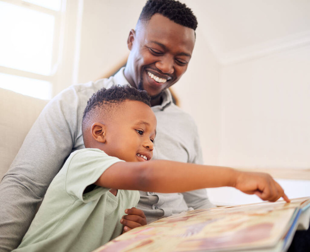Young happy cheerful African american father reading a book with his son sitting on the couch at home. Little boy enjoying a story and learning while sitting on the sofa with his dad. - Photo, Image