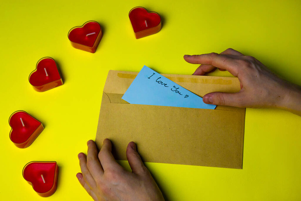 De hand van een vrouw stopt een blauw briefje in een kartonnen enveloppe met een handgeschreven tekst die IK HOUD VAN JULLIE voor haar minnaar op Valentijnsdag. Liefdesbrief op een gele achtergrond plat. Rode kaarsen in de vorm van harten. - Foto, afbeelding