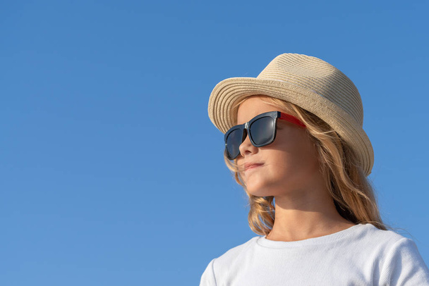 Close-up portrait of a young girl in sunglasses and a hat against the blue sky. View from a low angle. - 写真・画像