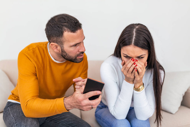 Infidelity. Jealous boyfriend Showing his Cheating girlfriend her Phone Demanding Explanation Sitting On Sofa Indoor. Wife caught by huband while cheating with mobile phone - 写真・画像