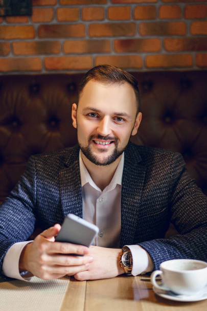 A young man with a neat beard dressed in a business suit with a smartphone and a cup of coffee at a table in a restaurant. A businessman with a phone and a cup of tea or coffee in a restaurant  - 写真・画像