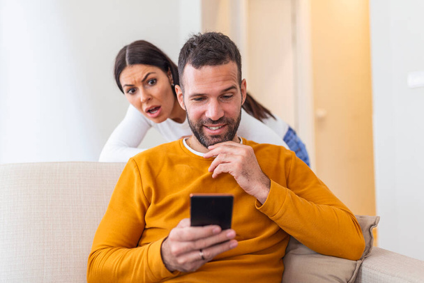 Pettäjä mies dating verkossa älypuhelimen ja tyttöystävä vakoilee istuu sohvalla kotona
 - Valokuva, kuva