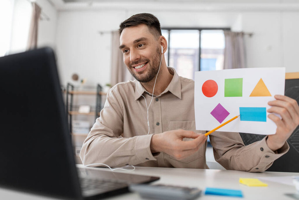distance education, primary school and remote job concept - happy smiling male teacher with laptop and picture of geometric shapes in different colors having online class at home office - Foto, afbeelding
