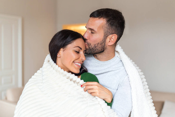 Romantic couple on a winter holiday. Man and woman standing together in a hotel room wrapped in blanket. Couple embracing and smiling. - Photo, Image
