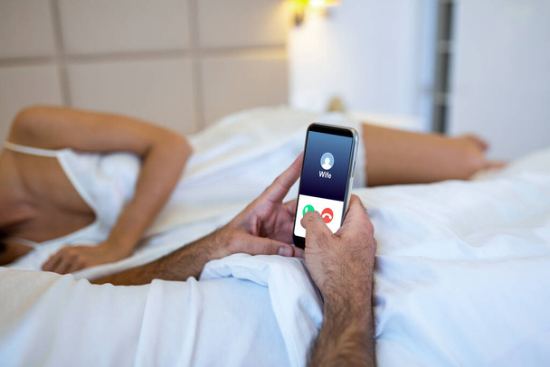 Cheating unfaithful man lying with mistress in hotel bed. Call from wife to mobile phone. Cheater having affair with secret lover and relationship with another woman. Infidelity and love triangle. - Photo, Image
