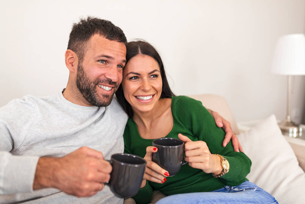 True love. Cheerful romantic couple is sitting on sofa, cozy and smiling. They are drinking tea, wearing casual clothes.Cheerful young couple in the morning at home. - Photo, Image