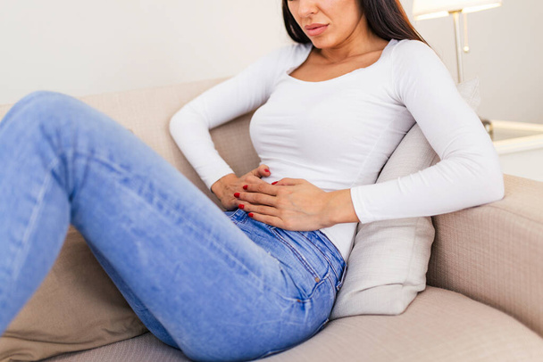 Woman in painful expression holding hands against belly suffering menstrual period pain, lying sad on home bed, having tummy cramp in female health concept - Foto, Imagen