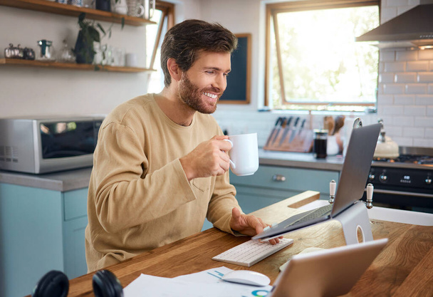 Happy young caucasian man drinking coffee while working on laptop in kitchen, checking his email or searching information while doing freelance work at home. Smiling young male using internet banking - 写真・画像
