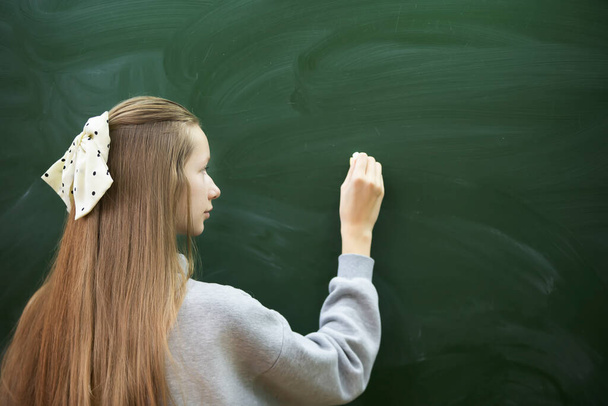 A teenage girl of middle school age writes with chalk on a blackboard. - Fotó, kép
