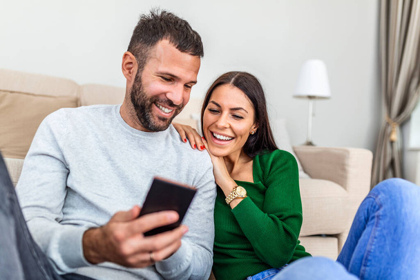 Smiling young couple sit relax on couch in living room have fun using smartphone together, happy Caucasian man and woman rest on sofa watch video on cellphone, make self-portrait picture on cell - Fotografie, Obrázek