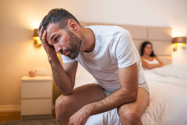 Junges Paar hat ein Problem. Guy sitzt auf dem Bett und schaut traurig weg, seine Freundin im Hintergrund. Verärgertes junges Paar hat Probleme mit Sex - Foto, Bild
