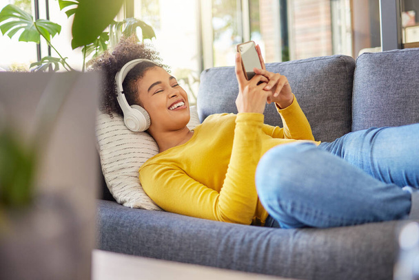 Jonge vrolijke gemengde ras vrouw het dragen van een koptelefoon en het luisteren naar muziek tijdens het typen van een bericht op een telefoon thuis. Een content Spaanse vrouw met een krullend afro met behulp van sociale media op een mobiele telefoon, terwijl - Foto, afbeelding