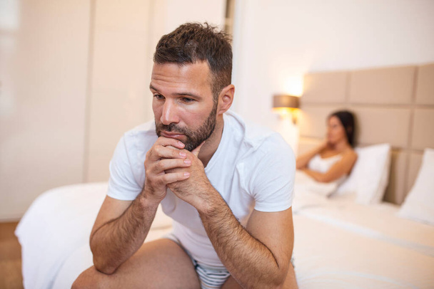 Young couple having a problem. Guy is sitting on bed and looking sadly away, his girlfriend in the background. Upset young couple having problems with sex - Photo, Image