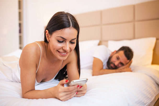 fiatal pár az ágyban férj frusztrált ideges és elégedetlen, míg az internet-függő felesége használja a mobiltelefon a szociális hálózat függőség figyelmen kívül hagyja őt kapcsolatban családi problémák - Fotó, kép