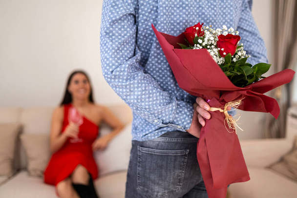 Arkasında gül buketi tutan erkek arkadaşına bakan mutlu bir kadın. Sevgililer Günü 'nde sevgilisine sürpriz yapmak için elinde çiçekler tutan yakışıklı adam. - Fotoğraf, Görsel