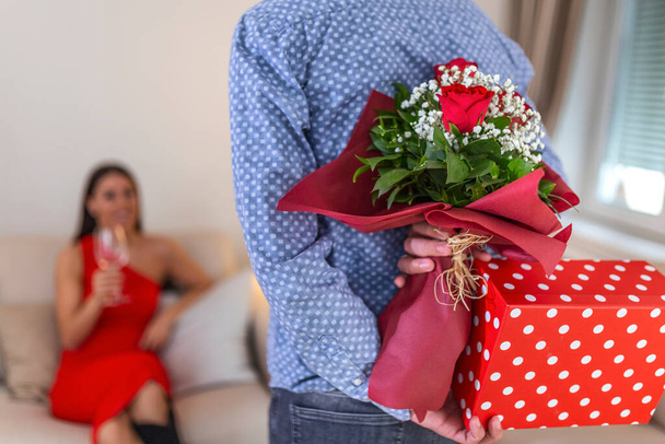 Unexpected moment in routine everyday life! Cropped photo of man's hands hiding holding chic bouquet of red roses and gift box behind back, happy woman is on blurred background - Foto, Bild