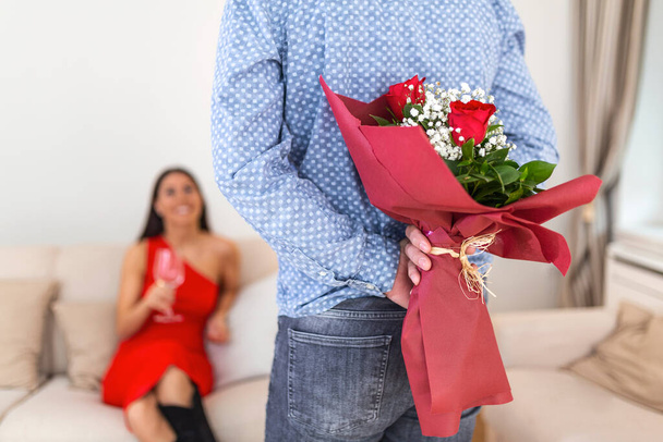 Счастливая женщина смотрит на своего парня, держащего букет роз за спиной. Мужчина держит цветы за спиной, чтобы удивить свою девушку, романтические счастливые пары на 8 марта счастливый женский день - Фото, изображение