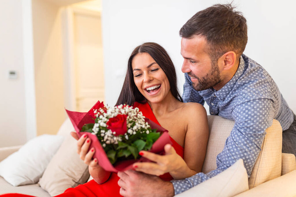 Excited young woman receiving unexpected Bouquet of red roses from husband at home, loving generous boyfriend making romantic surprise to attractive girlfriend on Valentines day occasion - Foto, imagen