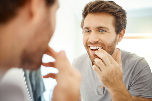 Yakışıklı bir adam evdeki banyoda dişlerini kontrol ediyor. Beyaz erkek, dişlerini temizliyor ve dairesindeki aynaya bakıyor. - Fotoğraf, Görsel