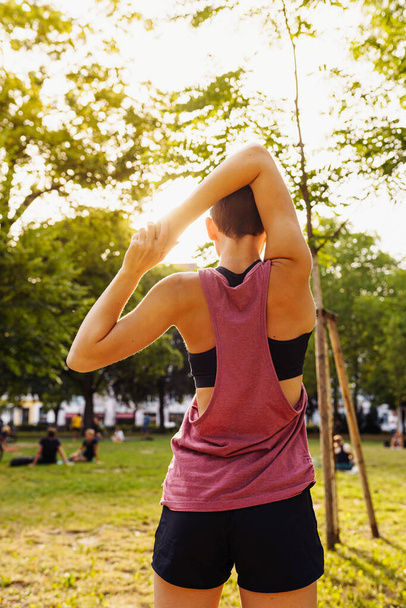 teenage girl in sports shorts and top does back stretch in public park in sun. Healthy active lifestyle, sports, outdoor fitness. Useful training, relaxation, muscle stretching. Rear view of sporty teenager girl practicing yoga in park at sunset.  - Fotoğraf, Görsel
