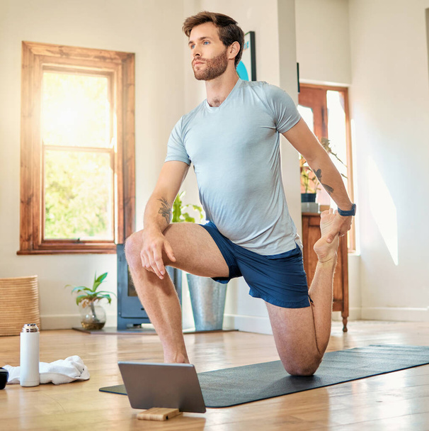 One fit young caucasian man stretching legs with lunge bodyweight exercise and holding foot while training with online tutorial on digital tablet at home. Guy gaining muscle, endurance, balance and - 写真・画像
