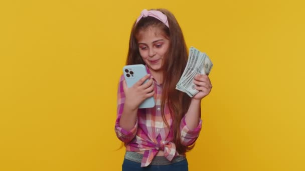 Smiling happy young child girl kid looking smartphone display sincerely rejoicing win, receiving money dollar cash banknotes, success lottery jackpot luck. Little toddler children on yellow background - Felvétel, videó