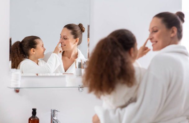 beauty, hygiene, morning and people concept - happy smiling mother and daughter looking to mirror at bathroom - Foto, afbeelding
