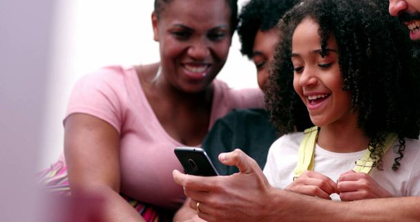 Счастливая многонациональная семья смеется и улыбается вместе глядя на телефон. Настоящие родители и дети смеются и улыбаются - Фото, изображение