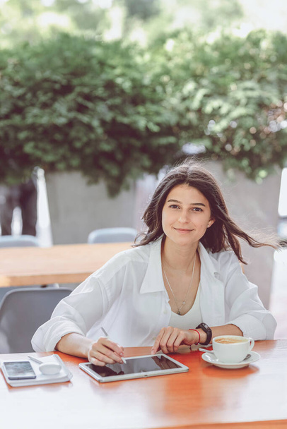 Caucasienne belle jeune blogueuse passionnée assise avec tablette numérique pour designer et tasse de café pendant les cours de langue dans un café sur la terrasse à l'extérieur. Concept d'emploi à distance et indépendant réussi - Photo, image