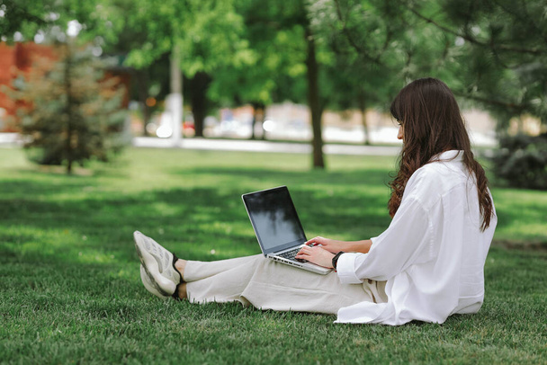 Beautiful Caucasian girl working with laptop computer sitting on green grass in summer park. Happy female outdoor working with technology, education and surfing the net, preparing for exams concept - Photo, Image