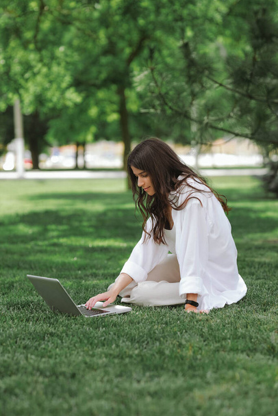 Yaz parkında yeşil çimlerde oturan güzel beyaz kız dizüstü bilgisayarla çalışıyor. Mutlu kadın dışarıda teknoloji, eğitim ve internette sörf yapıyor, sınav konseptine hazırlanıyor. - Fotoğraf, Görsel