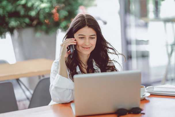 Портрет счастливого молодого офисного ассистента, работающего удаленно онлайн, сидящего с ноутбуком, планшетом и кофе на террасе, делающего заметки в повестке дня, в то же время разговаривающего с улыбкой по телефону   - Фото, изображение