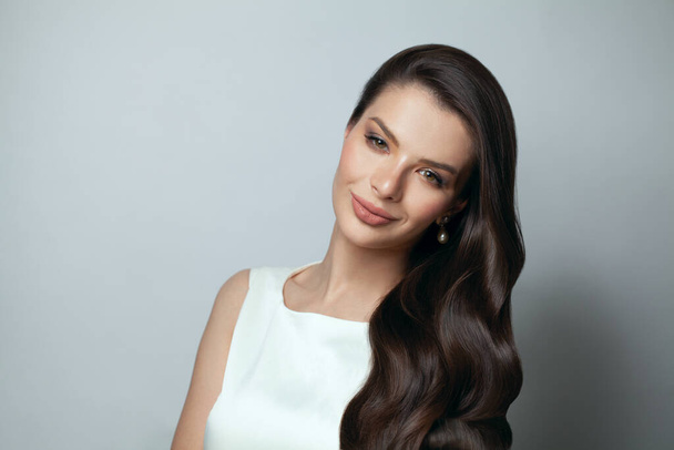 Mladý módní model brunetka s make-upem a zdravé dlouhé vlnité vlasy při pohledu na kameru a úsměv na šedém banneru pozadí. - Fotografie, Obrázek