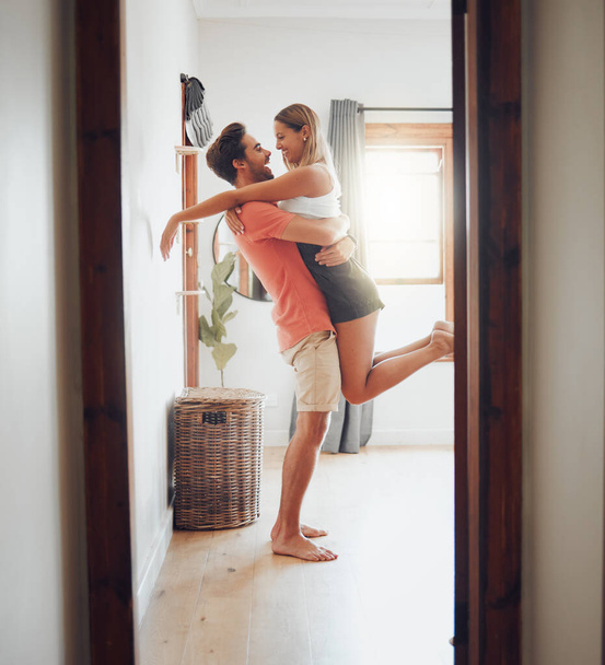 Happy young boyfriend holding girlfriend in arms as he lifts her up while they look into each others eyes and share intimate moment. Romantic young couple hugging and enjoying passionate dance at home - Photo, Image