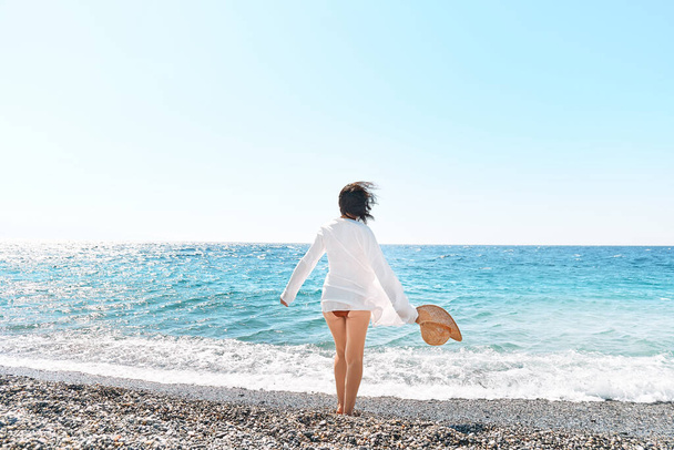 Zadní pohled bezstarostné ženy v slamáku a bílé košili kráčející po oblázkové pláži. Odpočinek a užít si dovolenou u moře v létě. Jednota s přírodou. Wellness, úspěch, svoboda a koncept cestování - Fotografie, Obrázek