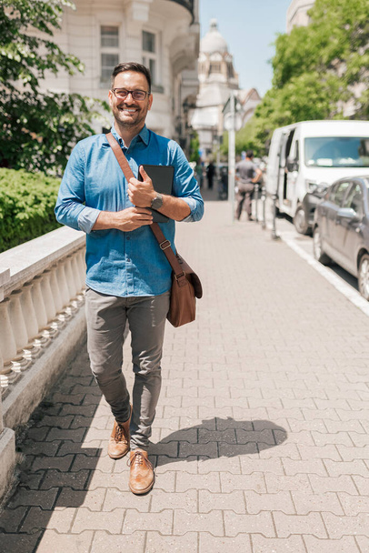 Usmívající se muž podnikatel s digitálním tabletem dívající se do kamery kráčející ulicí. Mladý profesionální dojíždějící muž nosí tašku na notebook. Chodí po chodníku ve městě.. - Fotografie, Obrázek