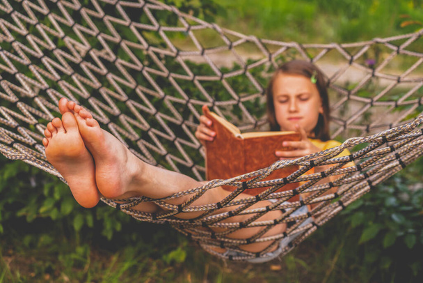 Young beautiful girl reads a book outdoors in a hammock.  Selective focus on feets. Horizontal image. - 写真・画像