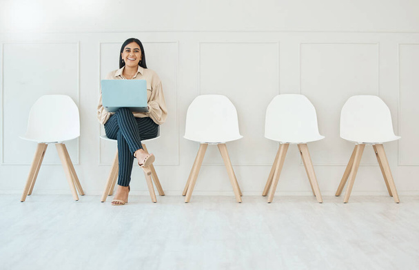 Fiatal boldog mosolygós kevert faj spanyol üzletasszony dolgozik laptop, miközben várja az interjút ül egy széken egy fehér fal egy irodában a munkahelyen. - Fotó, kép