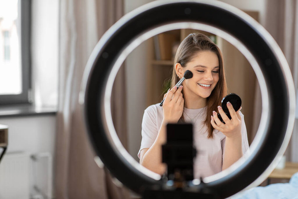 beauty blogging, technology and people concept - happy smiling girl blogger with ring light and smartphone applying make up at home - Foto, imagen