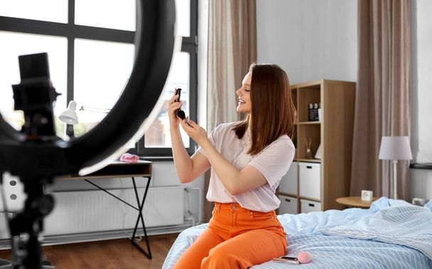 beauty blogging, technology and people concept - happy smiling girl blogger with ring light and smartphone applying make up at home - Foto, Imagem