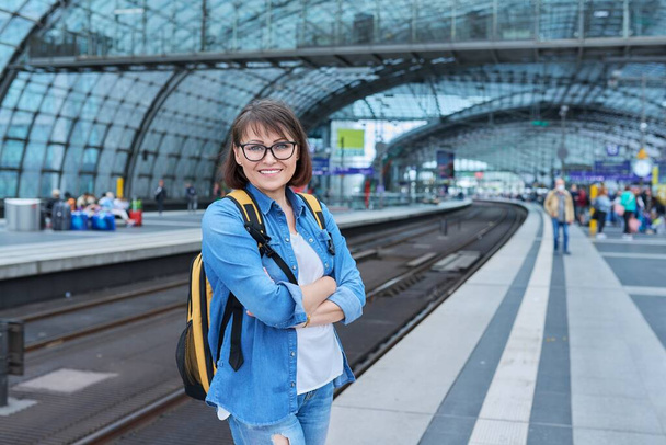 Woman passenger with backpack waiting on railway platform inside station, smiling female with crossed arms looking at camera. Rail transport, passenger transportation, journey, travel, trip, people - Photo, image