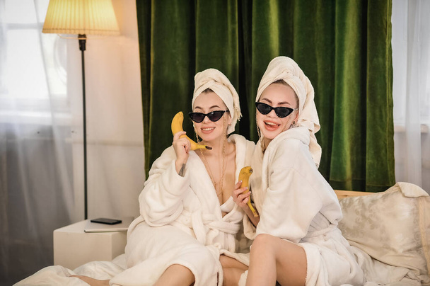 two beautiful women in bathrobes drinking juice and talking on mobile phone while sitting on sofa at home - Foto, immagini