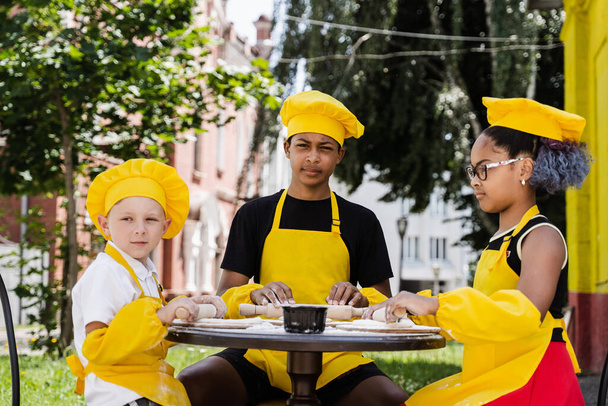 Multinational company of children cooks in yellow uniforms cooking dough for bakery. African teenager and black girl have fun with caucasian child boy and cook food - Foto, Imagem