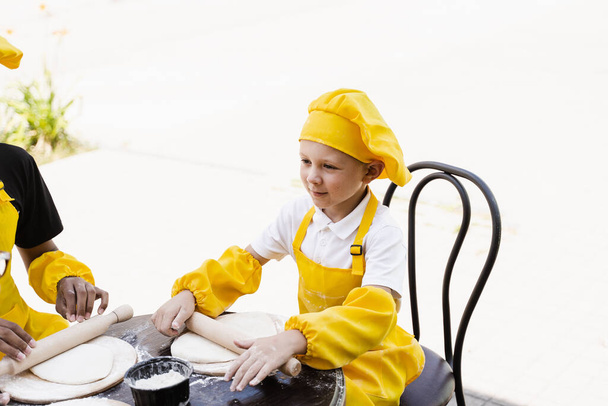 Handsome cook child in yellow chefs hat and apron yellow uniform rolling dough and cooking outdoor - 写真・画像