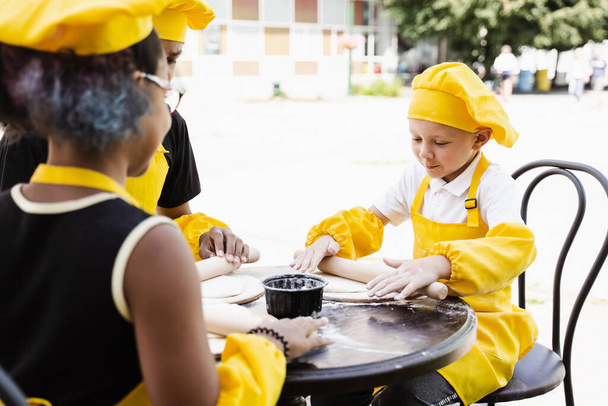 Handsome cook child in yellow chefs hat and apron yellow uniform rolling dough and cooking outdoor - Fotografie, Obrázek