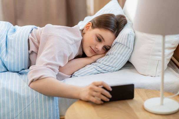 people, bedtime and rest concept - happy smiling teenage girl with smartphone lying in bed at home - Foto, Imagen