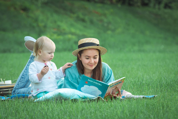 Mother is reading book for baby girl lying on the grass in the park. High quality photo - Zdjęcie, obraz