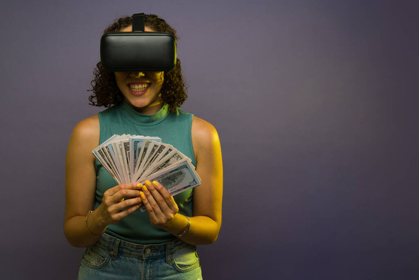 Cheerful woman in her 20s with virtual reality glasses earning a lot of money with virtual reality technology against a studio background - Foto, Bild