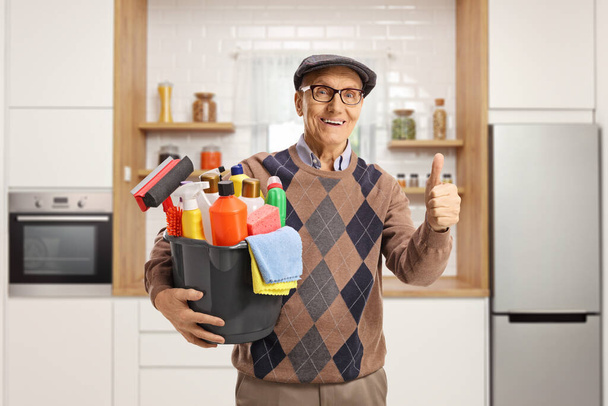 Elderly man holding a bucket with cleaning supplies and gesturing thumbs up inside a kitchen - Photo, image